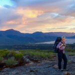Woman with large backpack stands in national park at sunrise with view of lake and mountains behind