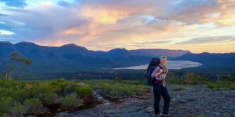 Woman with large backpack stands in national park at sunrise with view of lake and mountains behind